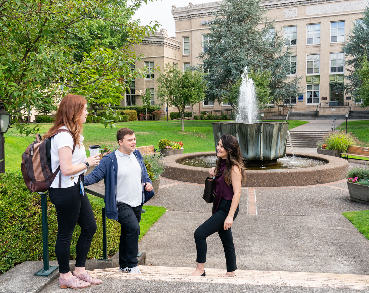 A young man and two young women in business casual attire talking on the steps in front of the Alumni Fountain.